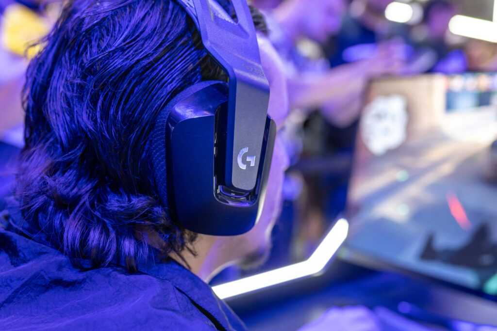 Focused gamer with headphones playing a video game on a PC under blue lighting in a crowded room.