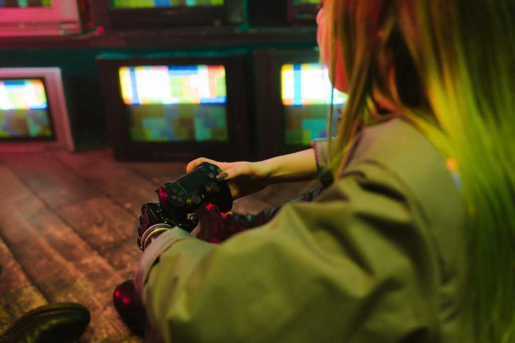 A woman with a prosthetic hand playing video games indoors amidst vintage TVs, showcasing innovation and leisure.